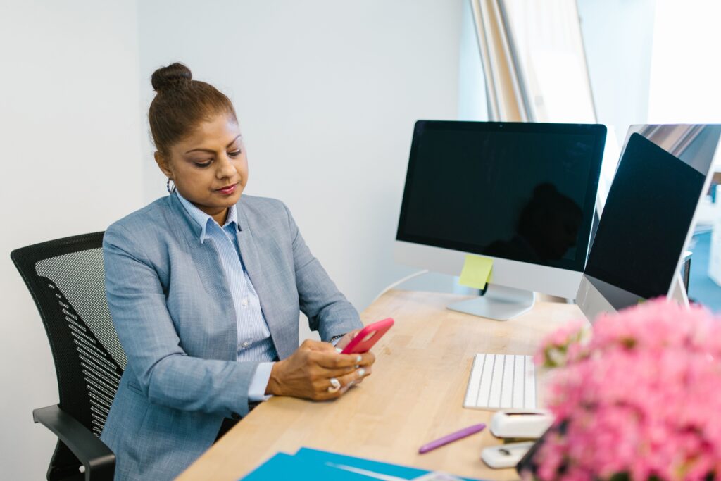 A businesswoman in a blue blazer uses her smartphone at a desk with computers and flowers.